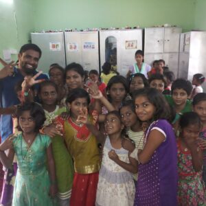A group of smiling children and a man pose together in a colorful classroom, making peace signs and cheerful gestures, reflecting a strong sense of community impact. Green walls, lockers, and bright natural light add to the lively scene.