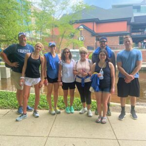 A group of eight people stand outdoors on a sidewalk near a small canal and lush greenery, smiling for the photo on a cloudy day. Dressed in athletic clothes and holding water bottles, they celebrate their community impact with buildings in the background.
