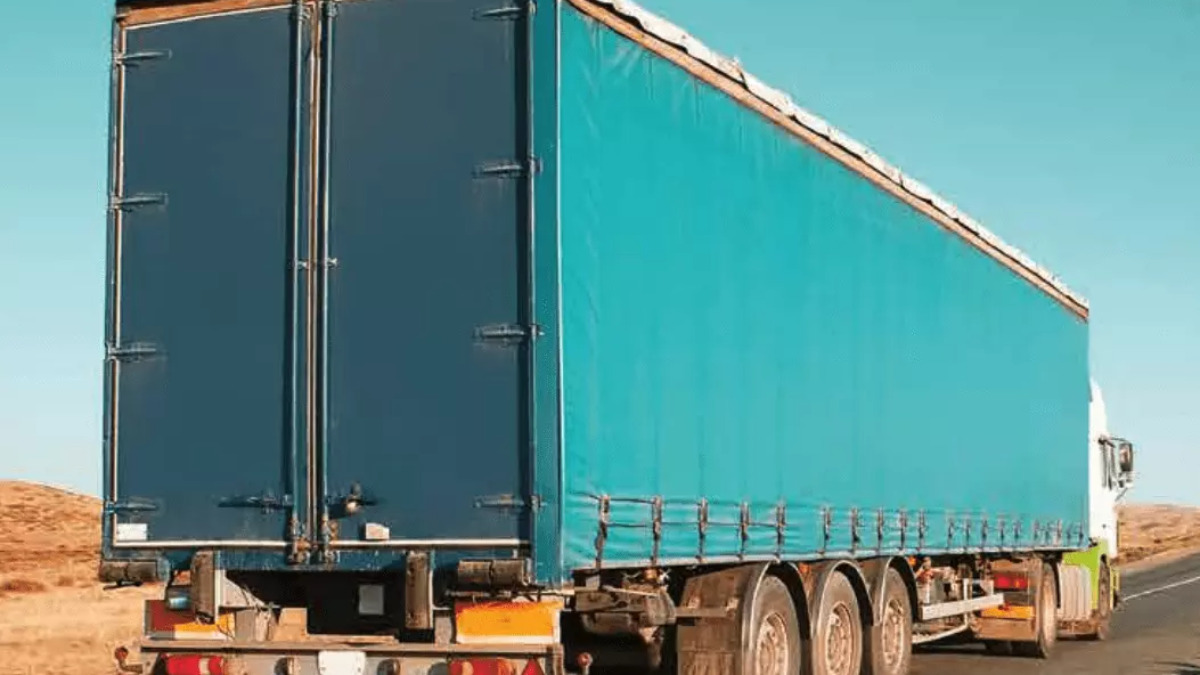 A large blue cargo truck with a covered trailer is driving on a paved road through a dry, open landscape under a clear blue sky.