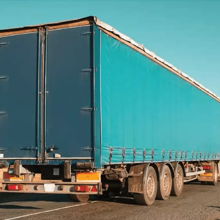 A large blue cargo truck with a covered trailer is driving on a paved road through a dry, open landscape under a clear blue sky.