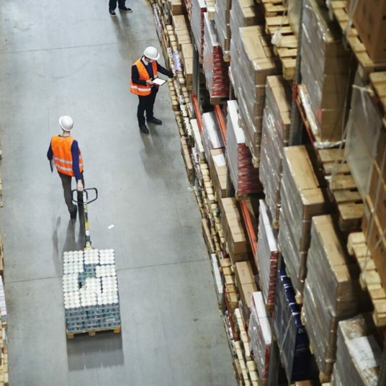Aerial view of two workers in safety vests and helmets in a warehouse aisle, one pulling a pallet jack with boxes, the other holding a clipboard among tall shelves filled with neatly stacked packages.