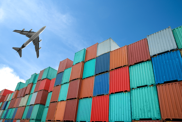 A commercial airplane flies above a large stack of colorful shipping containers under a clear blue sky, symbolizing global trade and transportation logistics.