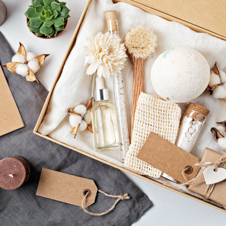 A gift box containing spa items such as a bath bomb, bar soap, glass bottles, dried flowers, and a loofah, surrounded by candles, cotton flowers, and blank brown gift tags on a fabric background.