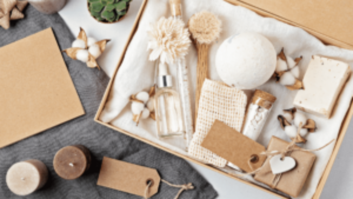 A gift box containing spa items such as a bath bomb, bar soap, glass bottles, dried flowers, and a loofah, surrounded by candles, cotton flowers, and blank brown gift tags on a fabric background.