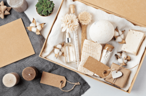 A gift box containing spa items such as a bath bomb, bar soap, glass bottles, dried flowers, and a loofah, surrounded by candles, cotton flowers, and blank brown gift tags on a fabric background.