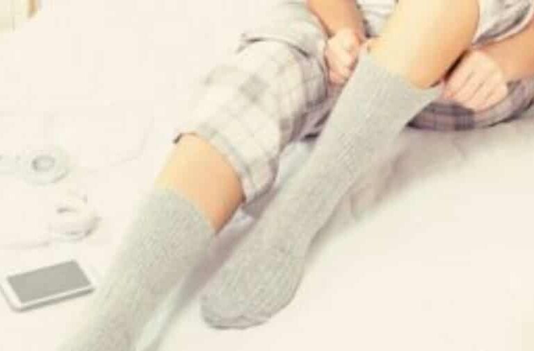 A person wearing plaid pajamas sits on a bed, pulling up gray socks. A smartphone and white headphones lie beside them.