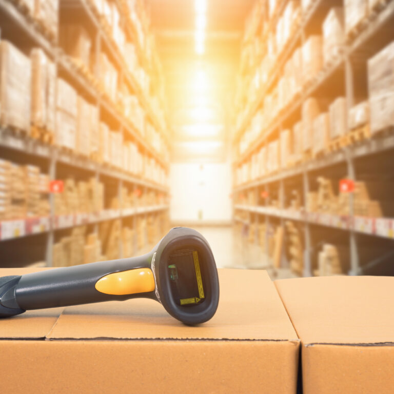 A barcode scanner resting on stacked cardboard boxes in a warehouse, surrounded by shelves filled with various boxes in the background, highlights efficient inventory management under bright sunlight.