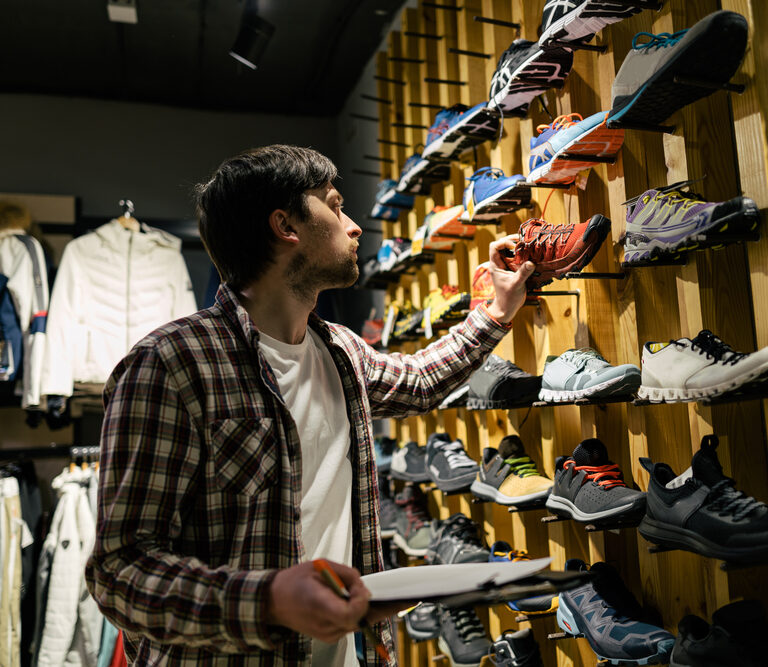 A man in a plaid shirt holds a clipboard and examines running shoes displayed on a wooden wall, focused on assortment planning in the sports store. Clothing and jackets hang neatly on racks in the background.