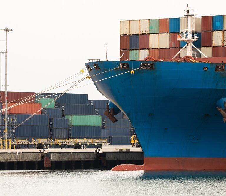 A large blue cargo ship docked at a port, with colorful shipping containers stacked both on the vessel and on the dock in the background.