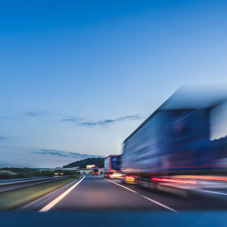 A blurred truck drives fast along a highway at dusk, with surrounding landscape and sky visible in the background. The image conveys a sense of speed and motion.