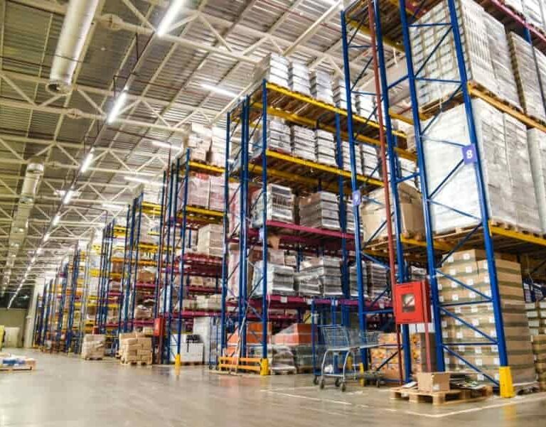 Large warehouse interior with tall blue and yellow shelving racks stacked with various pallets and boxes, organized in rows. The space is brightly lit with high ceilings and an orderly layout.