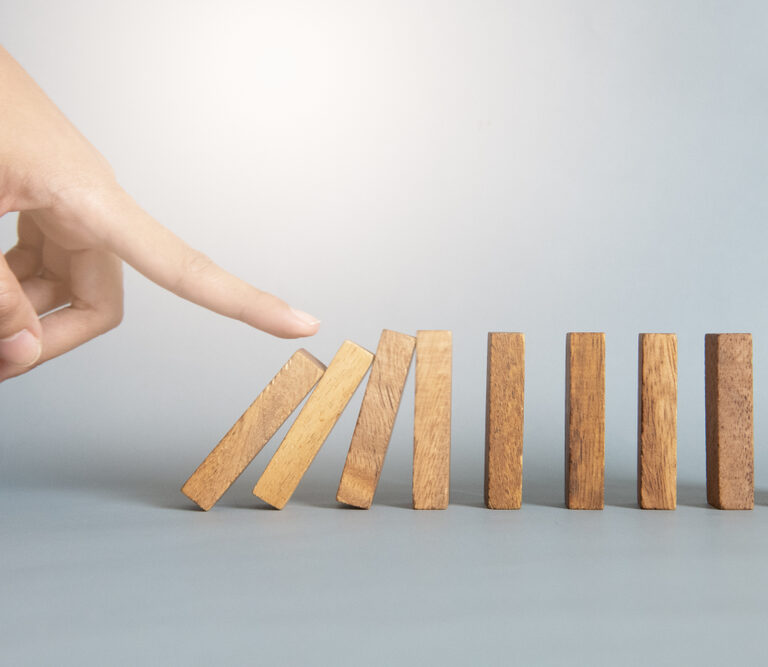 A hand is about to tip over the first of several upright wooden dominoes, illustrating a bullwhip effect chain reaction. The background is plain and light-colored.