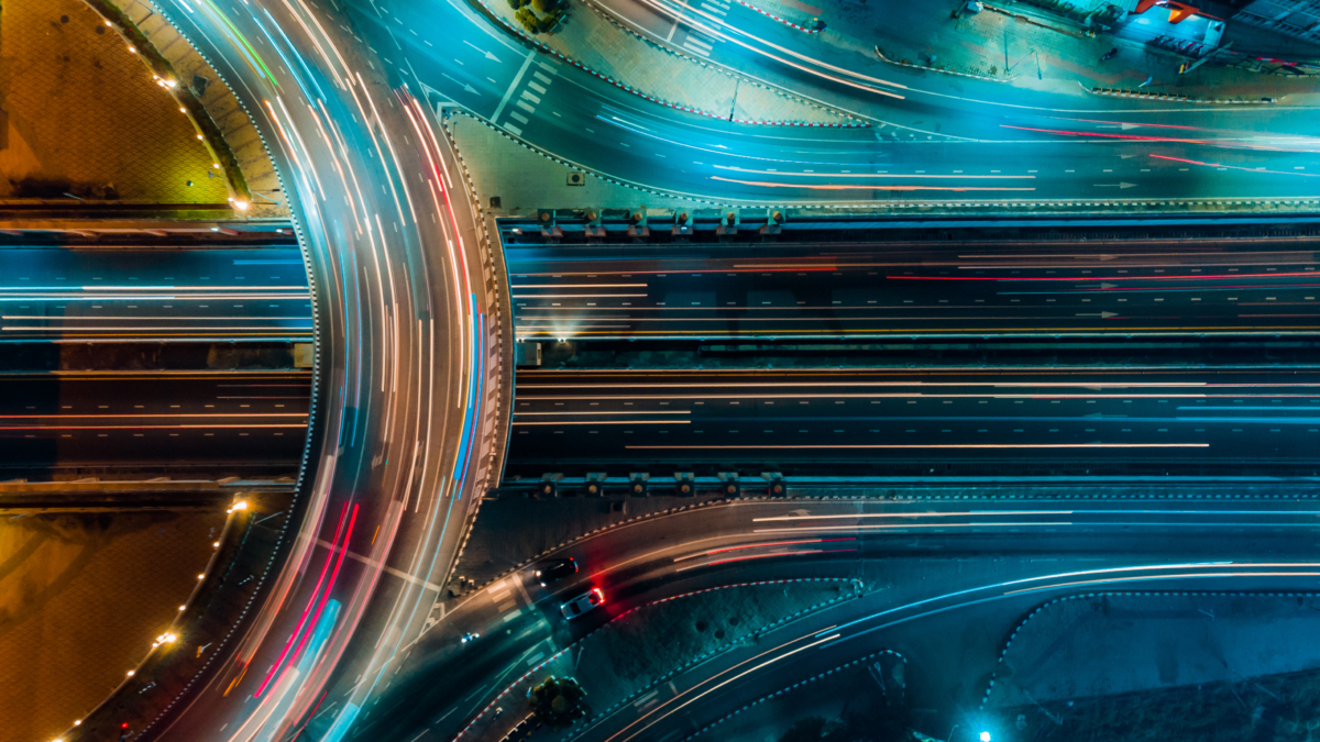 Aerial view of a brightly lit highway interchange at night, showing light trails from moving vehicles and curved roads intersecting with multiple lanes, surrounded by illuminated urban areas.