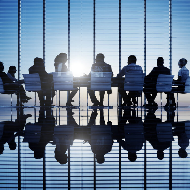A group of people in business attire sit around a conference table in a modern office with large windows, silhouetted against the sunlight, with their reflections visible on the shiny floor.