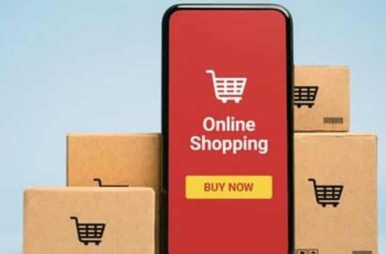 A smartphone displaying a red screen with a shopping cart icon, the words Online Shopping, and a Buy Now button, surrounded by several cardboard boxes with shopping cart symbols.