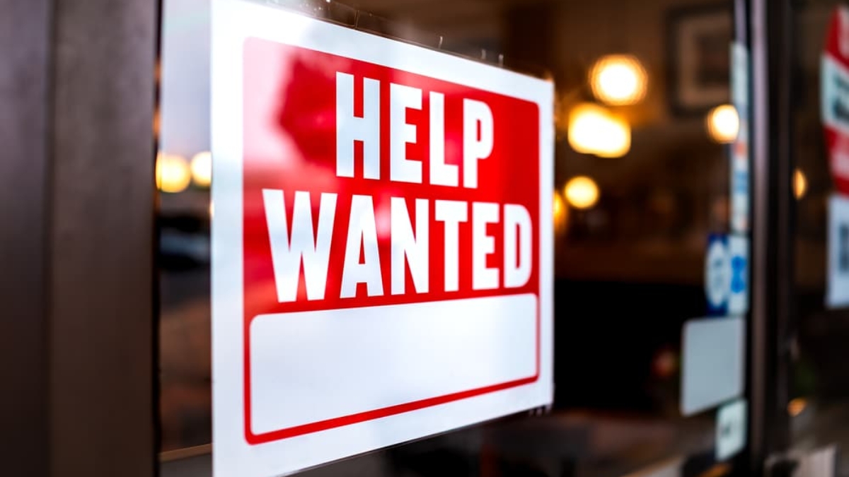 A red and white Help Wanted sign is displayed on a glass window or door, indicating that the business inside is hiring. The interior of the establishment is softly blurred in the background.
