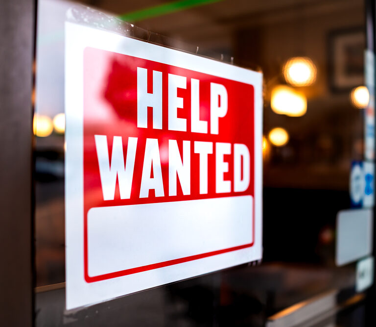 A red and white Help Wanted sign is displayed on a glass window or door, indicating that the business inside is hiring. The interior of the establishment is softly blurred in the background.