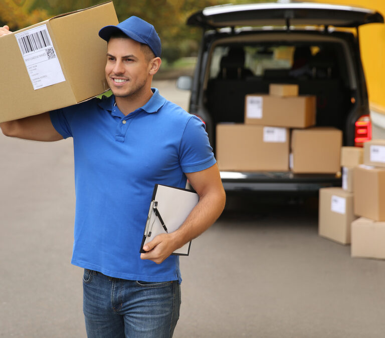 A delivery person in a blue uniform carries a large cardboard box on his shoulder and holds a clipboard, standing near a van with several packages loaded and stacked on the ground behind him.