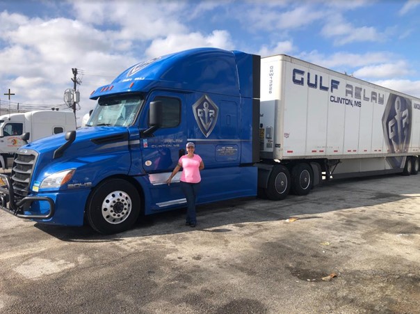 A woman in a pink shirt and cap stands beside a large blue semi-truck with Gulf Relay Clinton, MS written on the white trailer, parked in a lot under a partly cloudy sky.