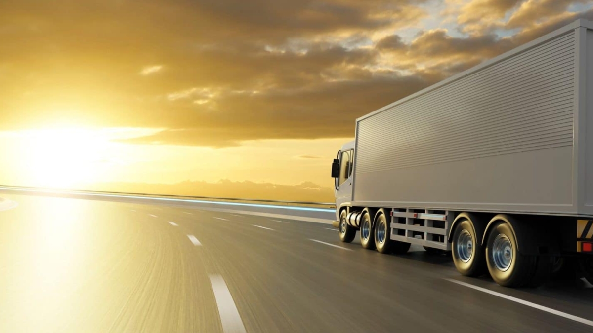 A large white semi-truck drives on a curved highway at sunset, with golden sunlight illuminating the sky and clouds, creating a dramatic and serene atmosphere.