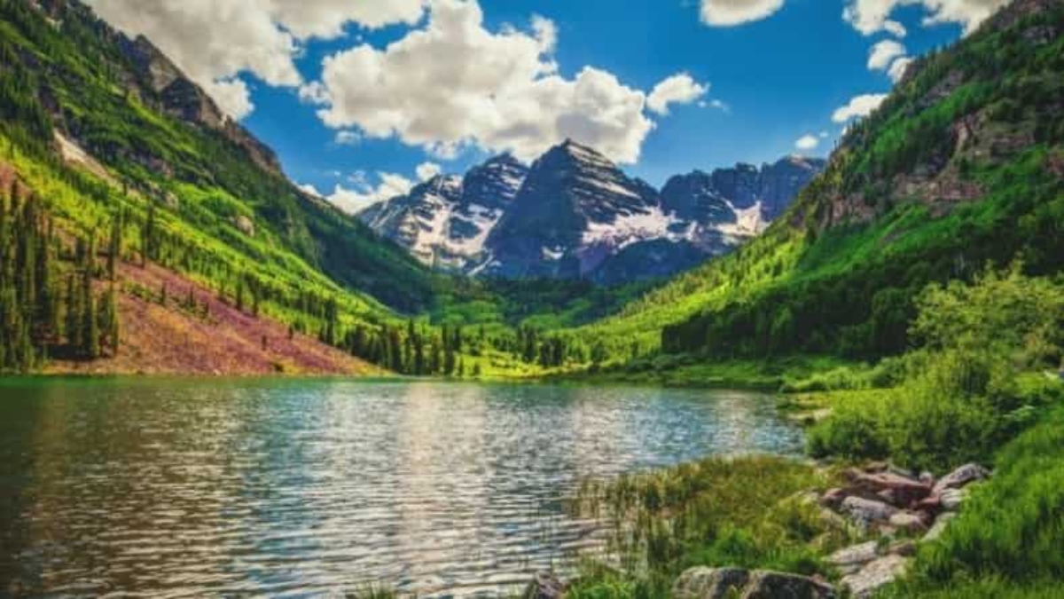 A serene mountain landscape with snow-capped peaks, lush green hillsides, and a clear lake reflecting the blue sky and clouds, surrounded by dense forest and rocky shore.
