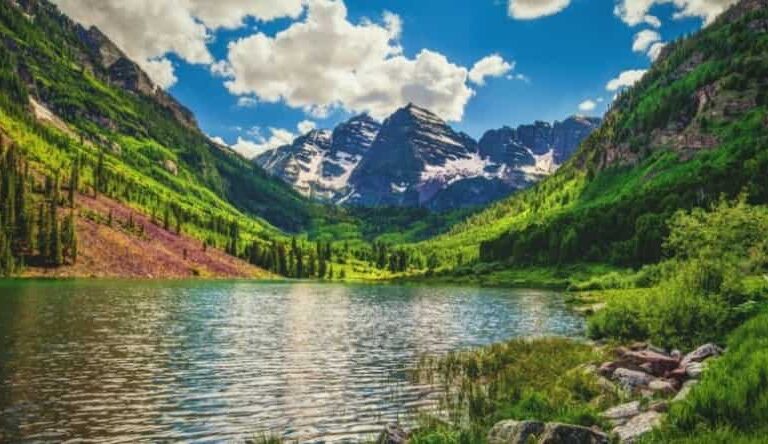A serene mountain landscape with snow-capped peaks, lush green hillsides, and a clear lake reflecting the blue sky and clouds, surrounded by dense forest and rocky shore.