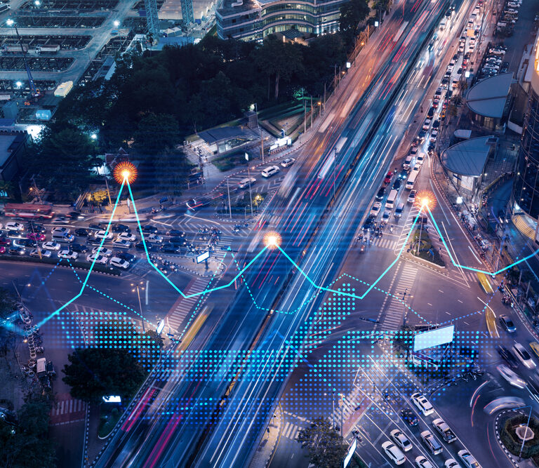 Aerial view of a busy city intersection at night with light trails from moving vehicles and digital data graphics, including a blue line graph and data points, overlaying the image to suggest smart city technology or traffic analytics.