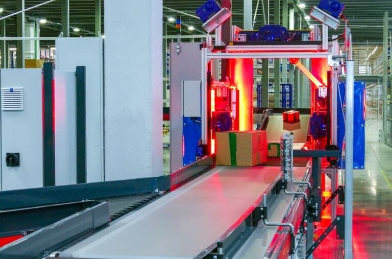 A conveyor belt in a warehouse moves packages through a scanning machine with red lights, surrounded by metal structures and shelving in an industrial setting.