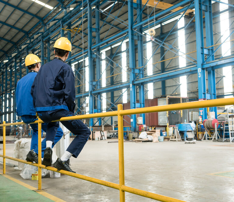 Two workers in blue uniforms and yellow hard hats sit on a yellow metal railing inside a spacious industrial warehouse with high ceilings and blue structural beams.