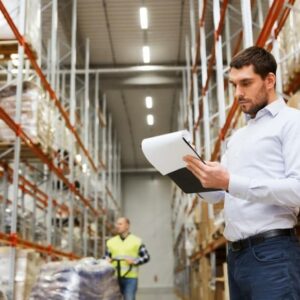 A man in a white shirt holds a clipboard and checks inventory in a large warehouse with tall shelves, while another worker in a safety vest walks in the background.