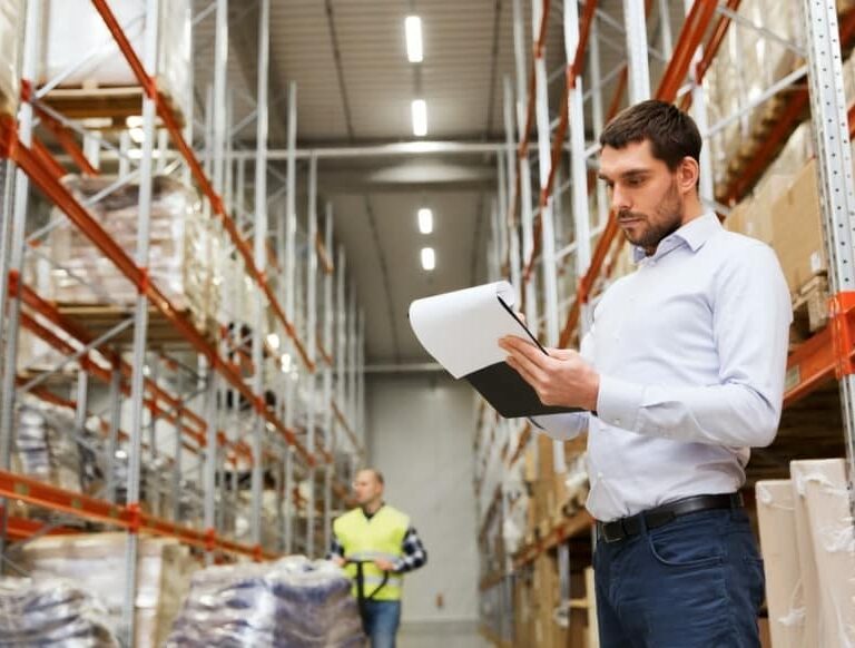 A man in a white shirt holds a clipboard and checks inventory in a large warehouse with tall shelves, while another worker in a safety vest walks in the background.