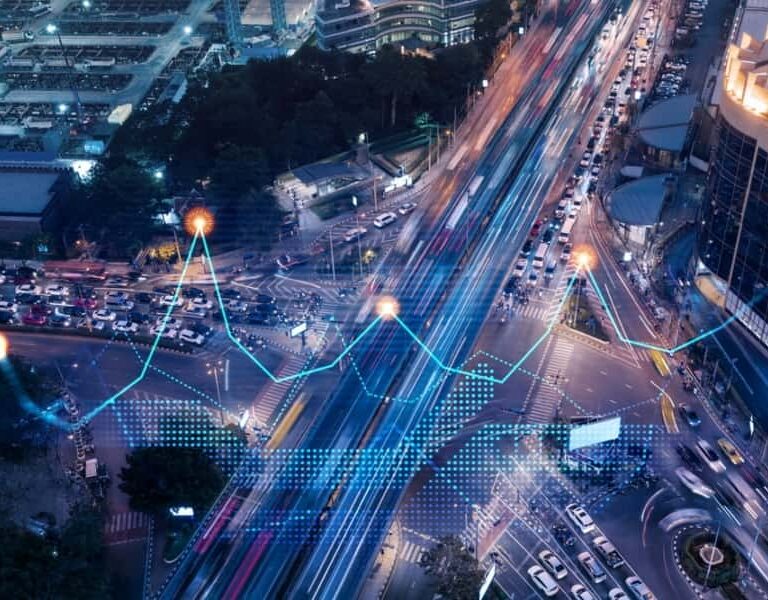A busy city intersection at night with light trails from cars. Digital blue and orange lines and data points overlay the scene, representing data analytics or smart city technology tracking traffic.