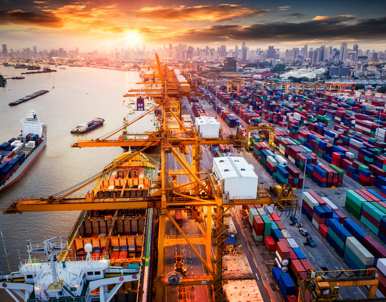 Aerial view of a busy shipping port at sunset, with large cranes, colorful stacked shipping containers, and cargo ships along the waterway, city skyline in the background.