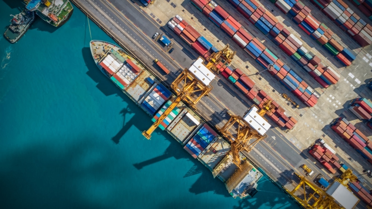 Aerial view of a busy shipping port with large cargo ships docked, yellow cranes loading containers, and rows of colorful shipping containers stacked along the dock—an essential hub in global supply chains beside turquoise water.