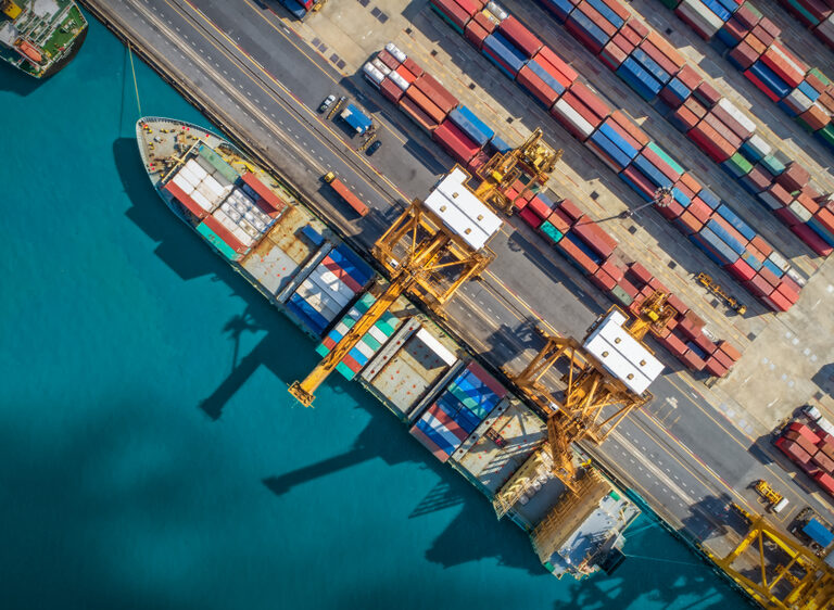 Aerial view of a busy shipping port with large cargo ships docked, yellow cranes loading containers, and rows of colorful shipping containers stacked along the dock—an essential hub in global supply chains beside turquoise water.