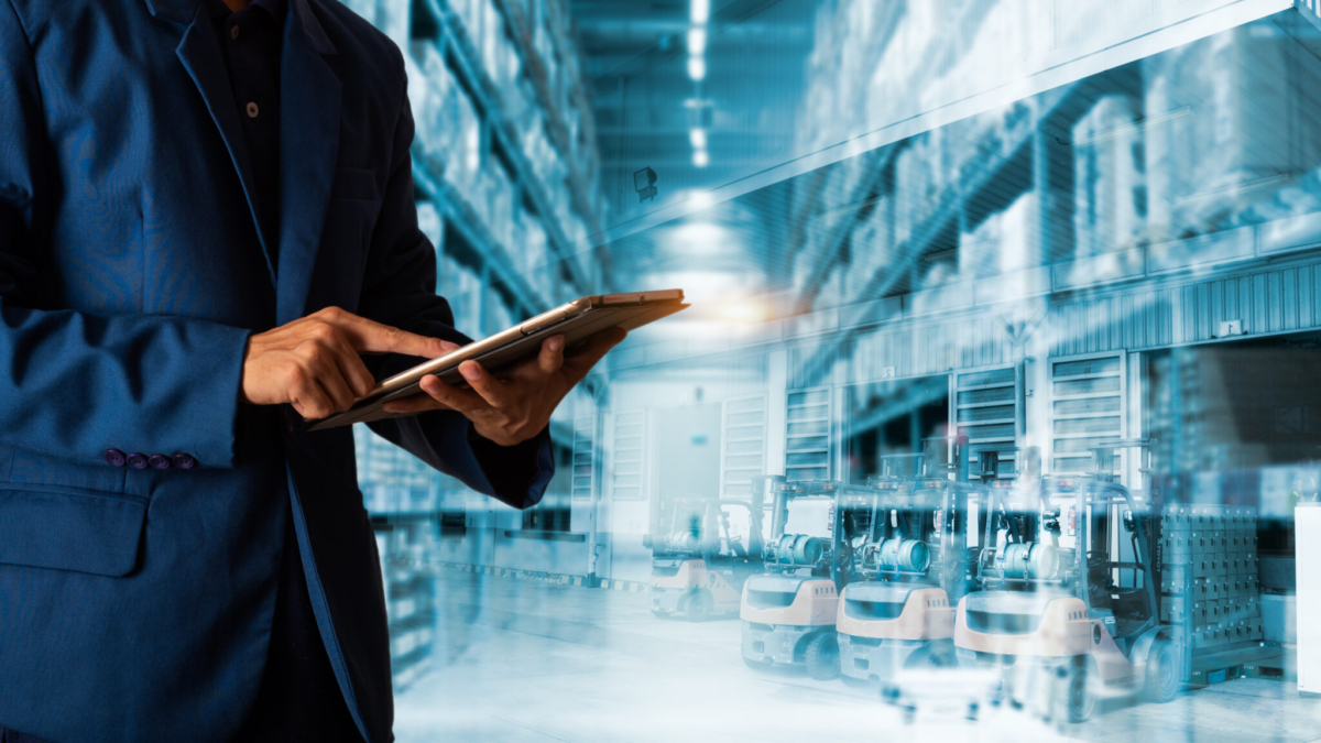 A person in a blue suit uses a tablet, standing in a warehouse with high shelves and rows of forklifts, representing modern inventory management and logistics technology.