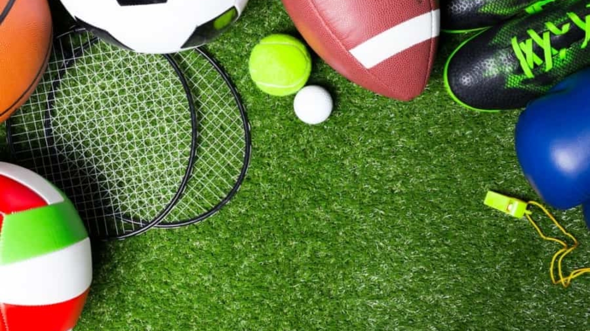Various sports equipment, including soccer ball, football, volleyball, tennis rackets, tennis ball, golf ball, green sports shoes, blue boxing glove, and a yellow whistle, arranged on green artificial grass.