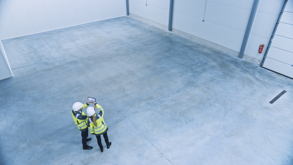 Two people in safety vests and helmets stand in an empty industrial warehouse, looking at a tablet as they discuss warehouse planning. The space features smooth concrete floors, white walls, and high ceilings.