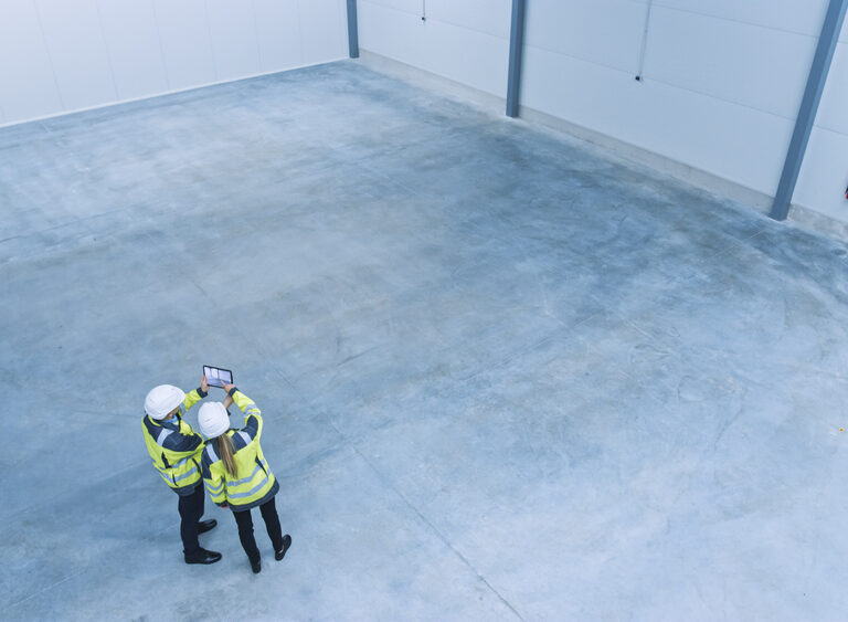 Two people in safety vests and helmets stand in an empty industrial warehouse, looking at a tablet as they discuss warehouse planning. The space features smooth concrete floors, white walls, and high ceilings.