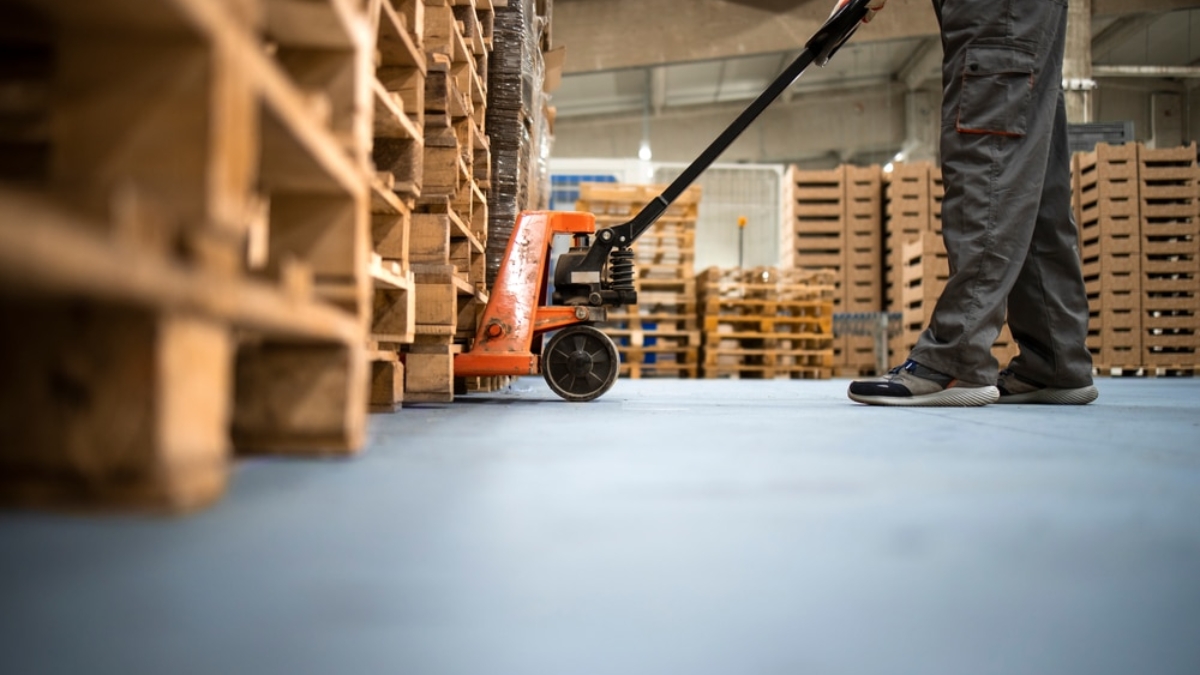 A worker in gray pants and sneakers operates an orange pallet jack, moving wooden pallets inside a warehouse with stacks of pallets in the background.