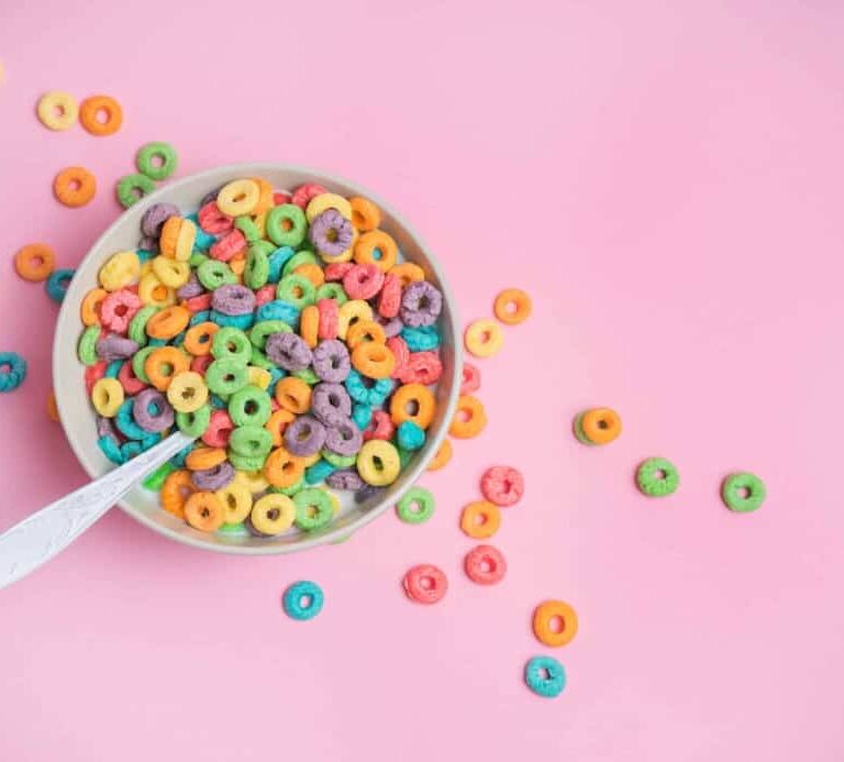 A bowl of colorful, round fruit-flavored cereal with a spoon sits on a pink background, with several cereal pieces scattered around the bowl.