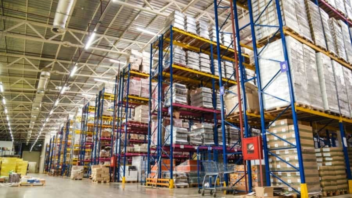 Large warehouse interior with tall blue and yellow metal shelving units stacked with pallets and boxes. Concrete floor, high ceilings with bright lighting, and some equipment and carts are visible.