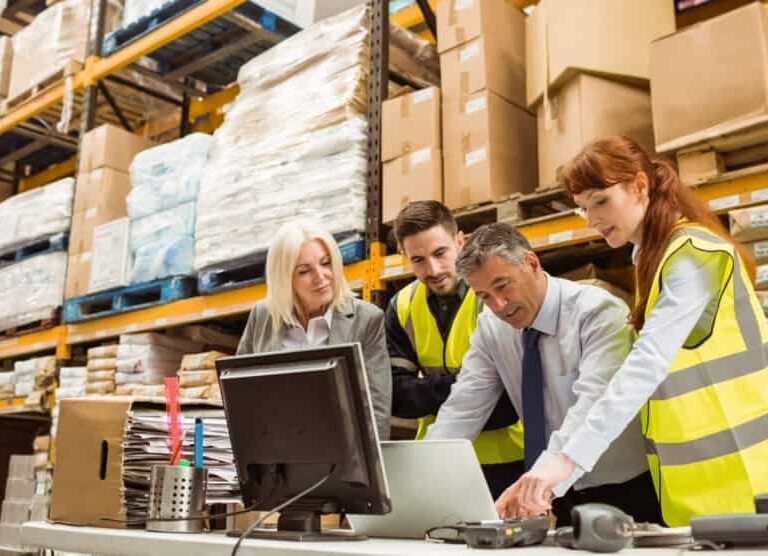 Four warehouse workers, three wearing yellow safety vests, gather around a desk with a computer and paperwork, discussing something in a busy storage facility with shelves stacked high with boxes and packages.