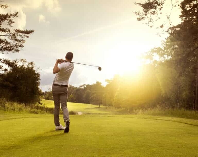 A person swings a golf club on a grassy course at sunset, surrounded by trees and a bright sky.