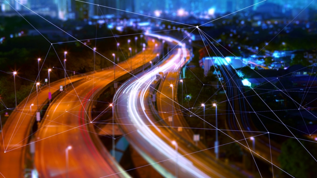 Blurred night view of a city highway with light trails from cars, overlaid with digital network lines suggesting connectivity, smart technology, and modern urban infrastructure.