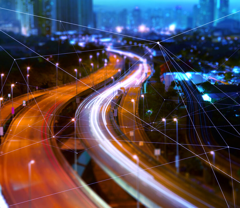 Blurred night view of a city highway with light trails from cars, overlaid with digital network lines suggesting connectivity, smart technology, and modern urban infrastructure.