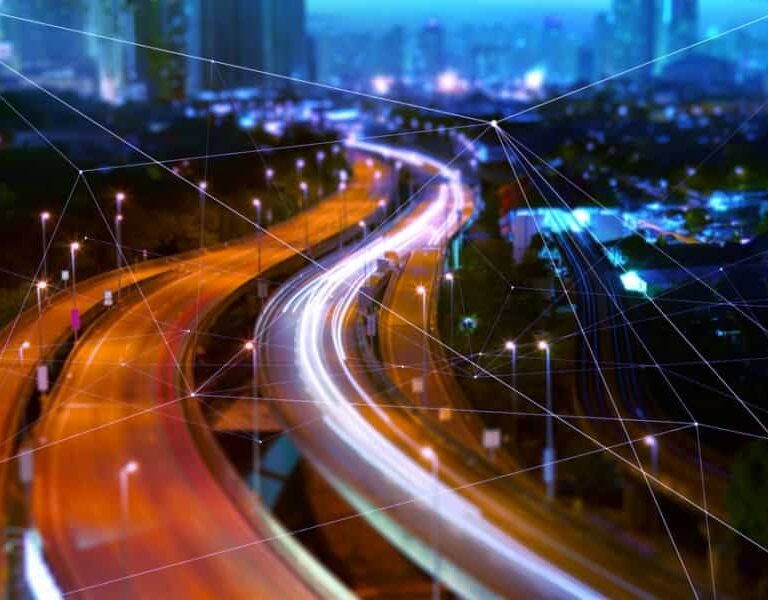 A city highway at night with bright, colorful light trails from moving vehicles. Skyscrapers rise in the background as translucent digital lines and nodes overlay the scene, hinting at a connected future powered by a Transportation Management System.