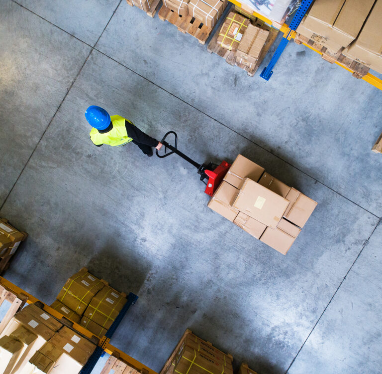 A warehouse worker wearing a blue helmet and yellow vest moves a pallet of boxes with a manual pallet jack, surrounded by stacks of cardboard boxes on pallets, viewed from above.