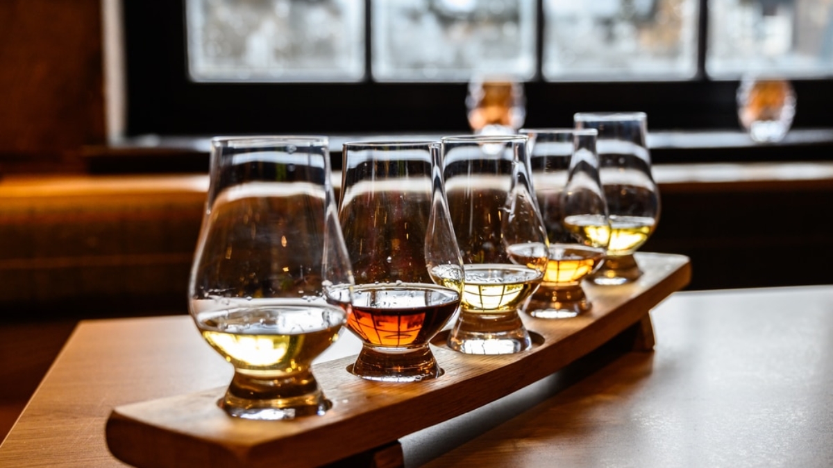 A wooden tray holds five glasses of whiskey in varying shades of amber, arranged in a row on a table, with a blurred window and cozy indoor setting in the background.