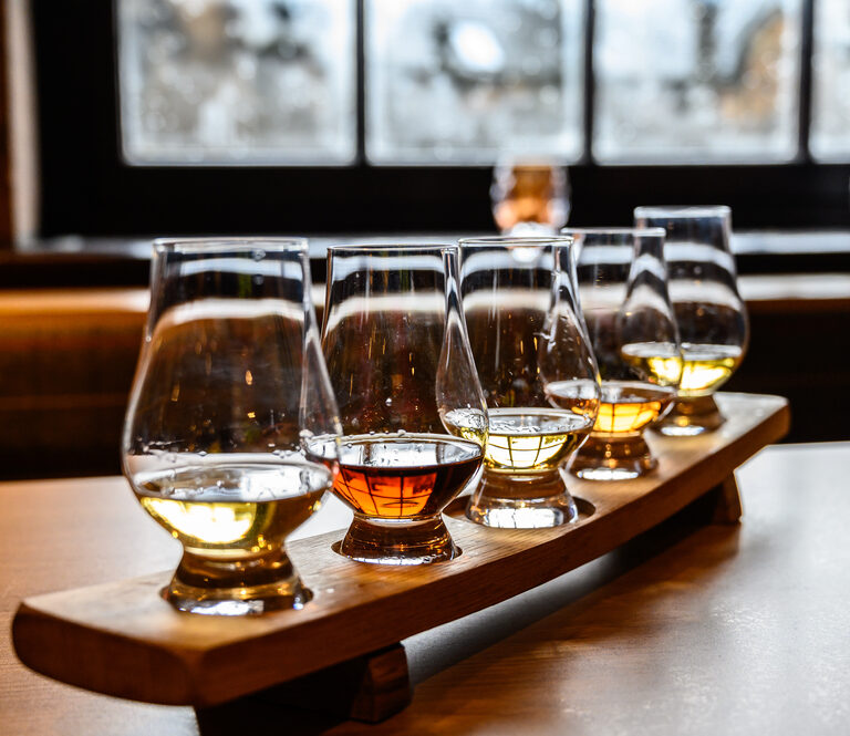 A wooden tray holds five glasses of whiskey in varying shades of amber, arranged in a row on a table, with a blurred window and cozy indoor setting in the background.